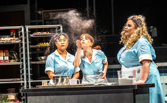 Three women wearing light blue dresses and white aprons stand in a kitchen.